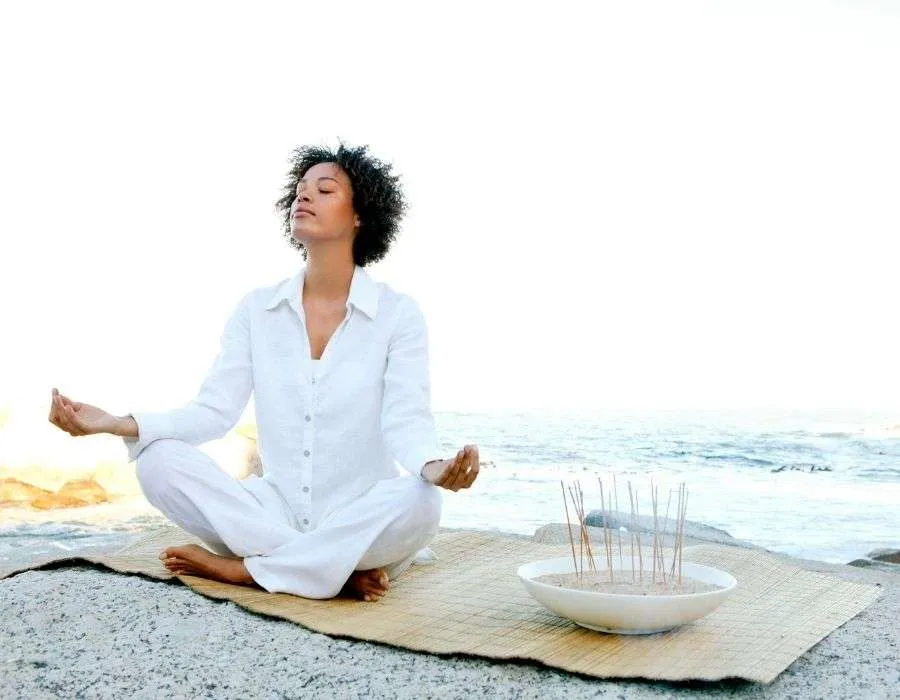 woman-in-all-white-on-rock-at-beach-meditating-sea-behind-benefits-of-meditation-blog-post