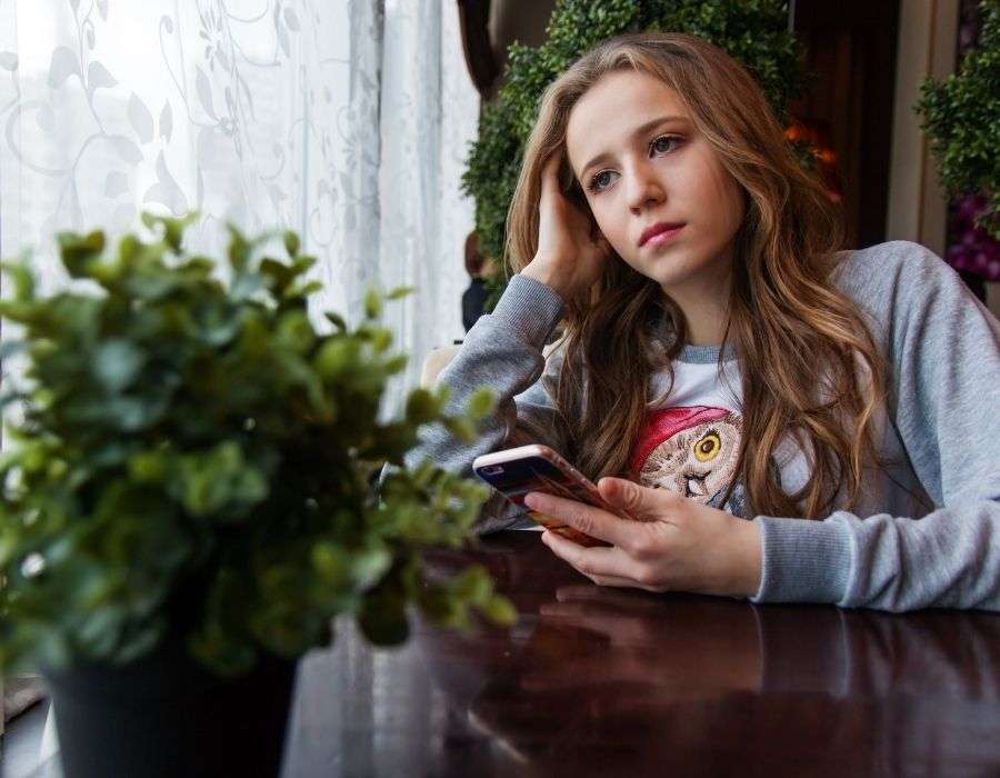 young-woman-long-brown-hair-sitting-at-table-by-window-plant-on-brown-table-hand-on-face-staring-blankly-bored-expression