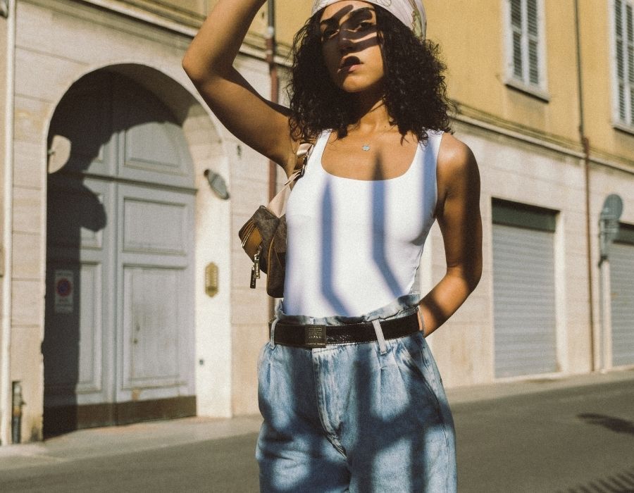 woman-dark-curly-hair-jeans-white-tank-top-standing-on-historic-street-hand-covering-sun-on-face