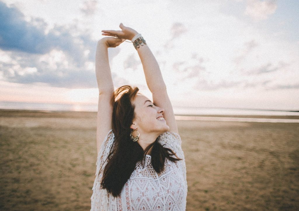 woman-long-dark-hair-white-shirt-on-beach-arms-stretched-above-head-happy-expression-know-your-story-blog-post